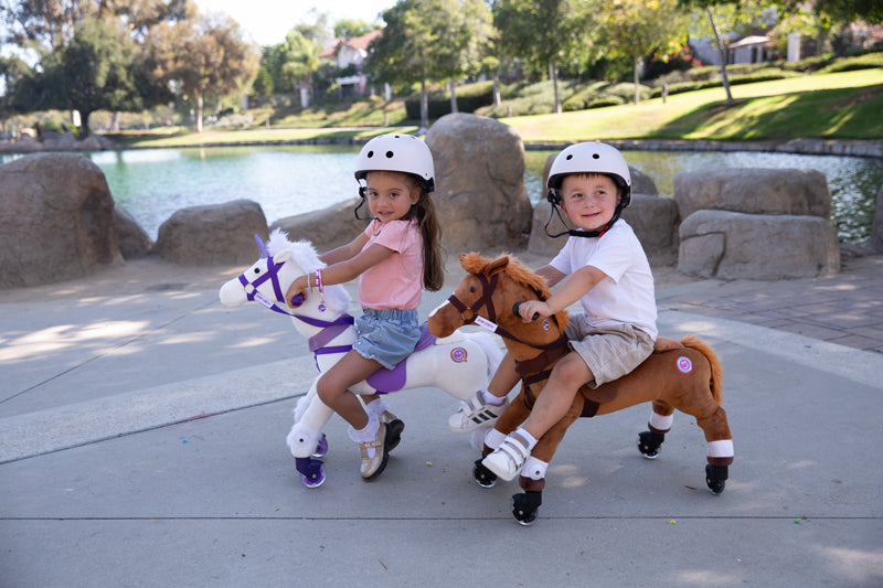 Two kids riding Power Ponies by a lake, both wearing helmets, enjoying a sunny day outdoors.