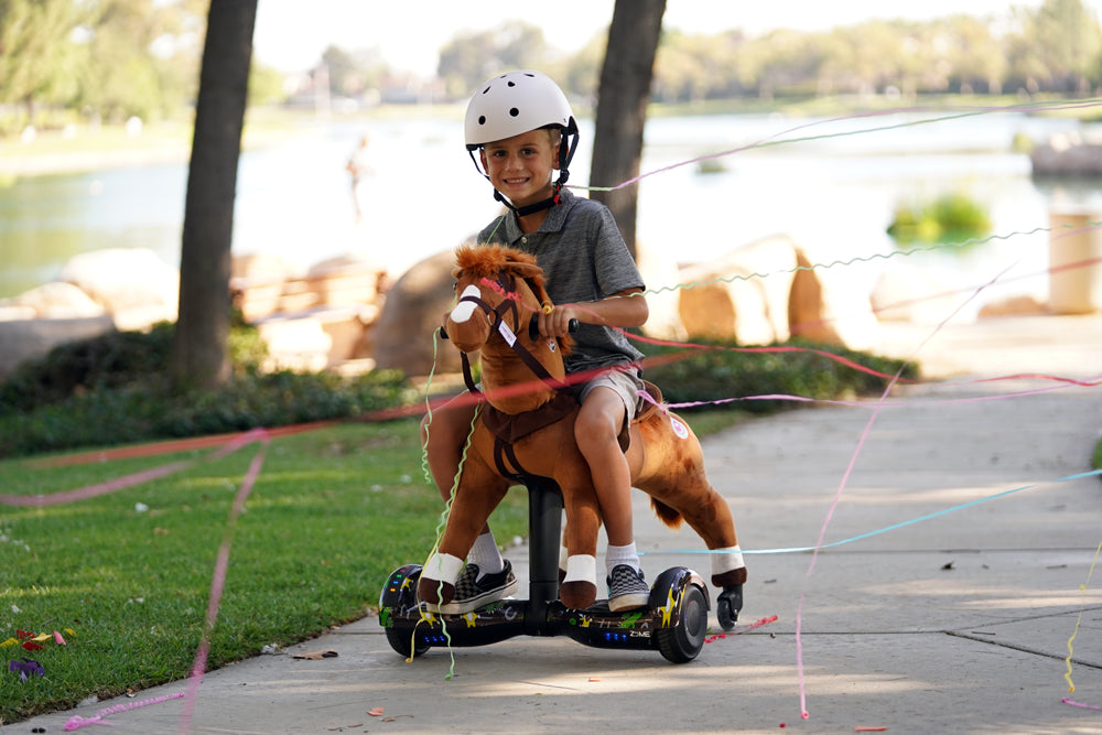 Smiling boy wearing a helmet riding a brown Power Pony on a lakeside path with colorful streamers.