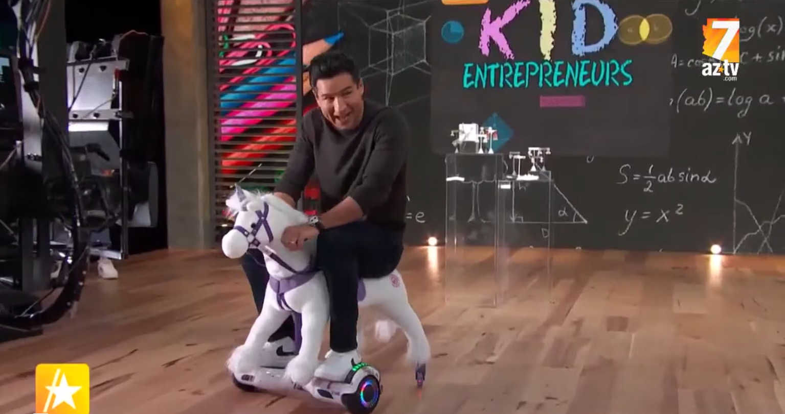 Mario Lopez riding a white Power Pony unicorn on set during a segment of Access Hollywood on kid entrepreneurs.